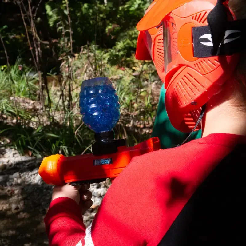 Person wearing red helmet, holding an orange gel blaster, aiming into greenery.