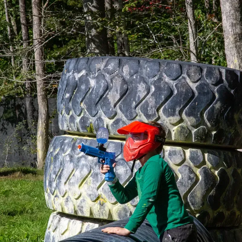 Person in red mask and green shirt holding a paintball gun, crouching behind a stack of tires in a wooded area.