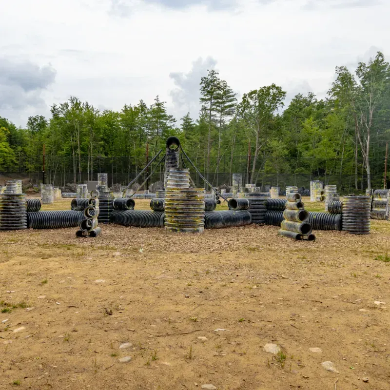 Paintball course with various bunkers on a sandy ground, forest in the background under a cloudy sky.