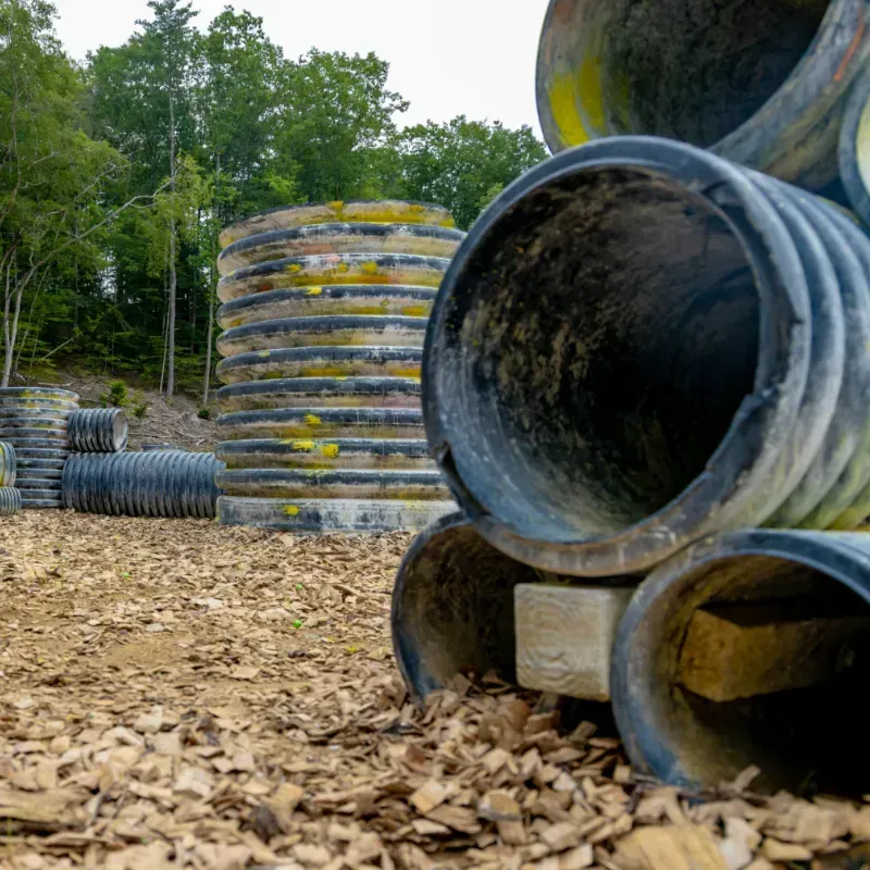 Paintball course with stacked corrugated pipes, a central tower, and a wooded background.