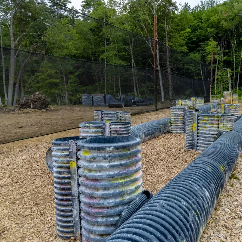 Paintball course with corrugated pipes and barrels, set in a wooded area.