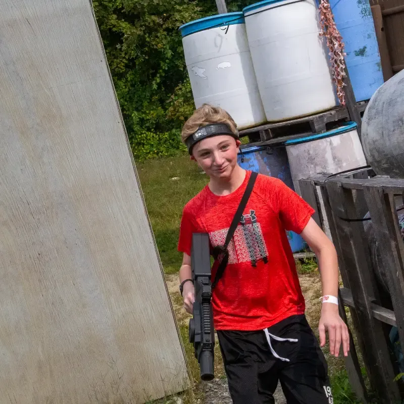Boy in red shirt and headband holds a toy gun, smiling in a paintball arena.