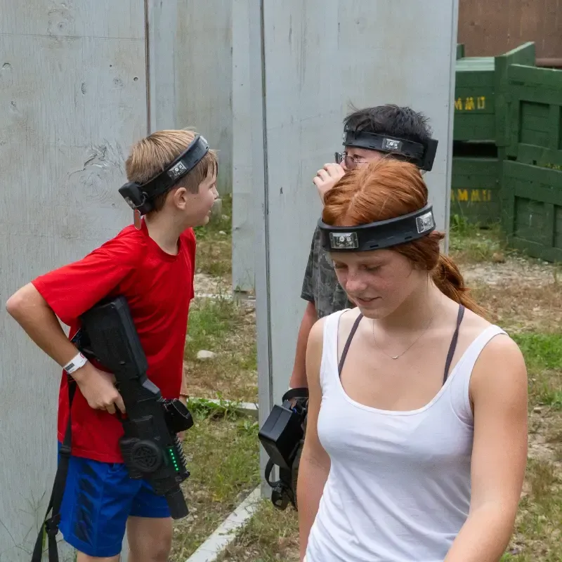 Three teens playing laser tag outdoors, holding guns, wearing headbands, near wooden walls.