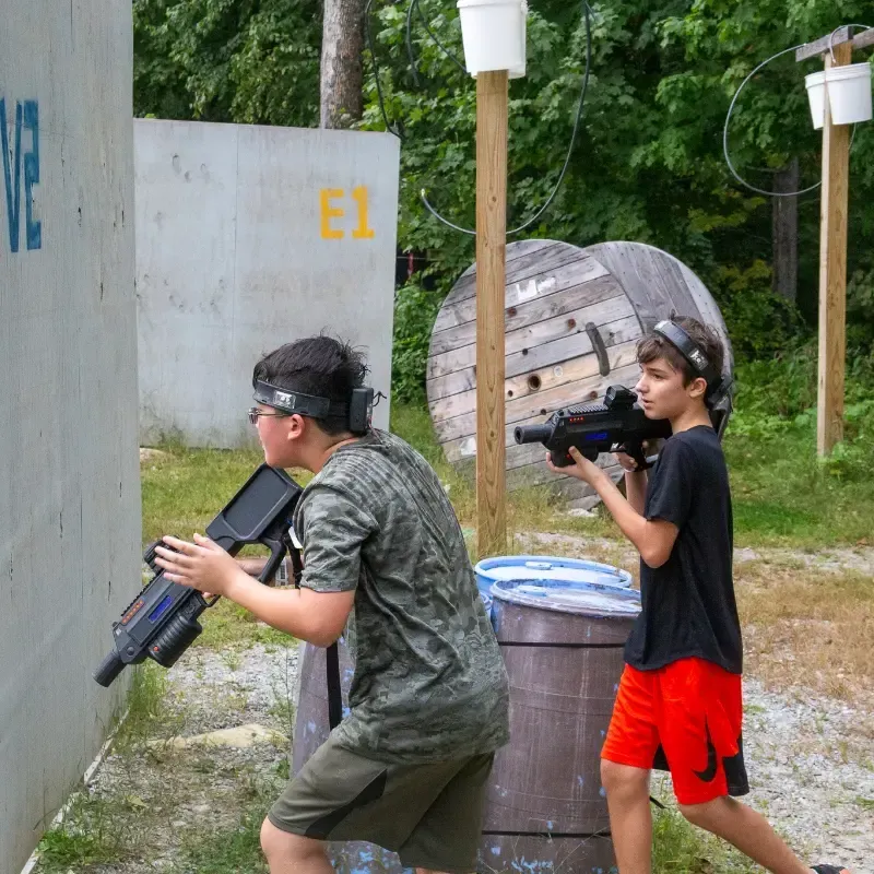 Two kids playing laser tag outdoors, aiming guns. One hides behind a wall, the other moves forward.