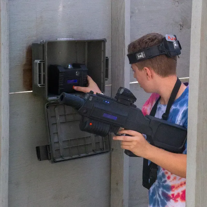 A young person in a tie-dye shirt holding a laser tag gun retrieves a module from a wall-mounted box.
