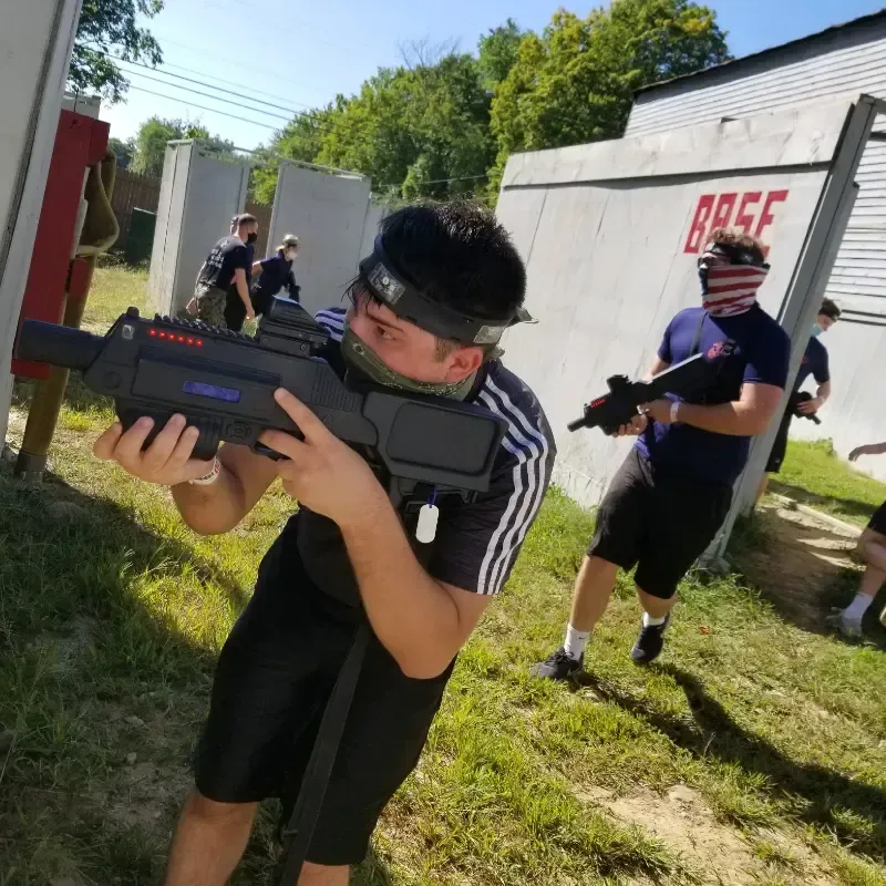 Group playing laser tag outdoors, aiming toy guns. One wears a headband and holds a large gun. 