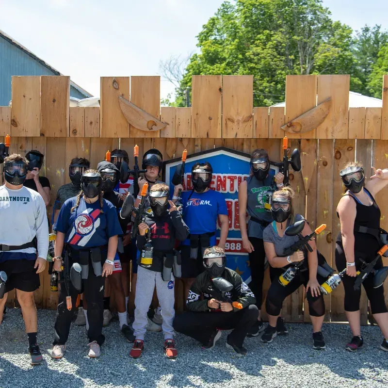 Group of people in paintball gear posing in front of a wooden barrier, holding paintball guns.