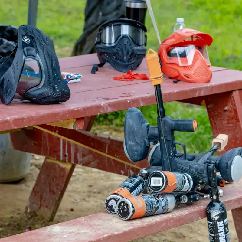 Paintball gear on a red picnic table: masks, gun, orange tank, and paint bottles.