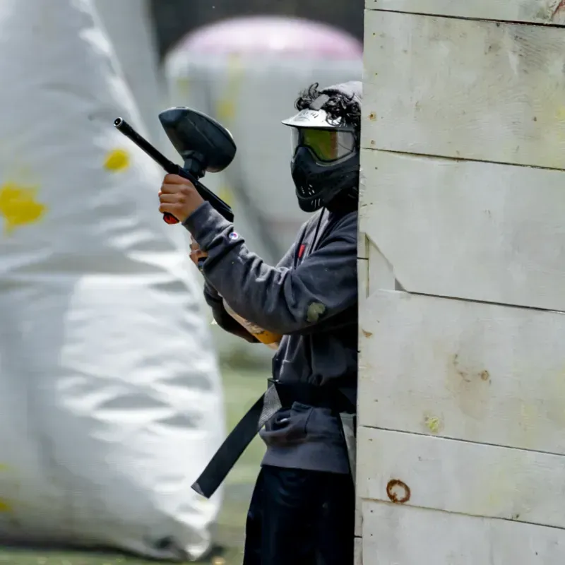 Person playing paintball, aiming gun from behind a white wooden barrier. Wearing a mask and grey clothing.