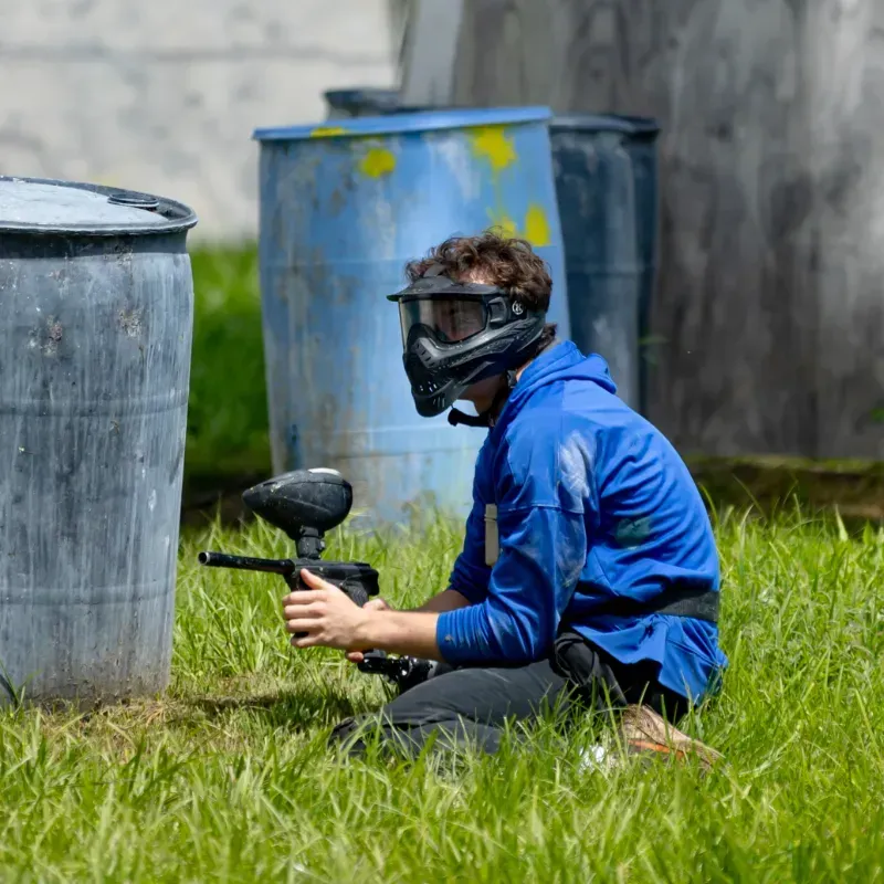 Person in blue crouching, playing paintball, aiming gun near blue barrels on a grassy field.