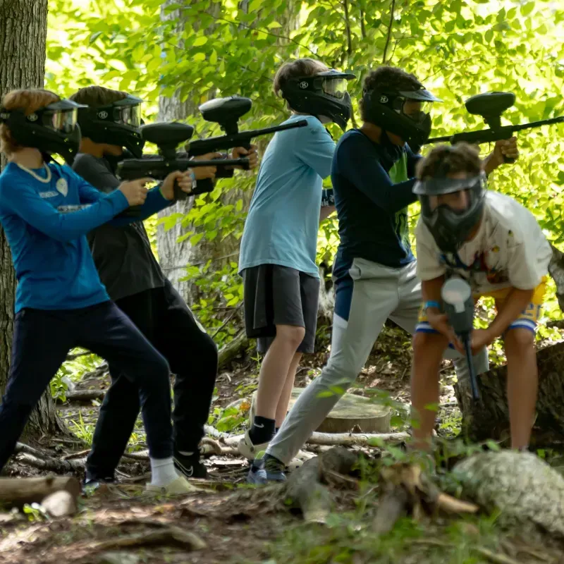 People playing paintball in a wooded area, aiming markers while wearing protective masks.