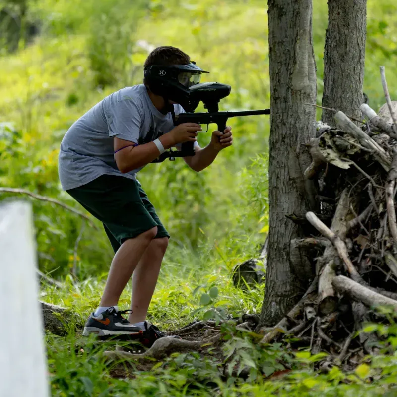 Person in paintball gear aims a marker behind a tree in a wooded area.