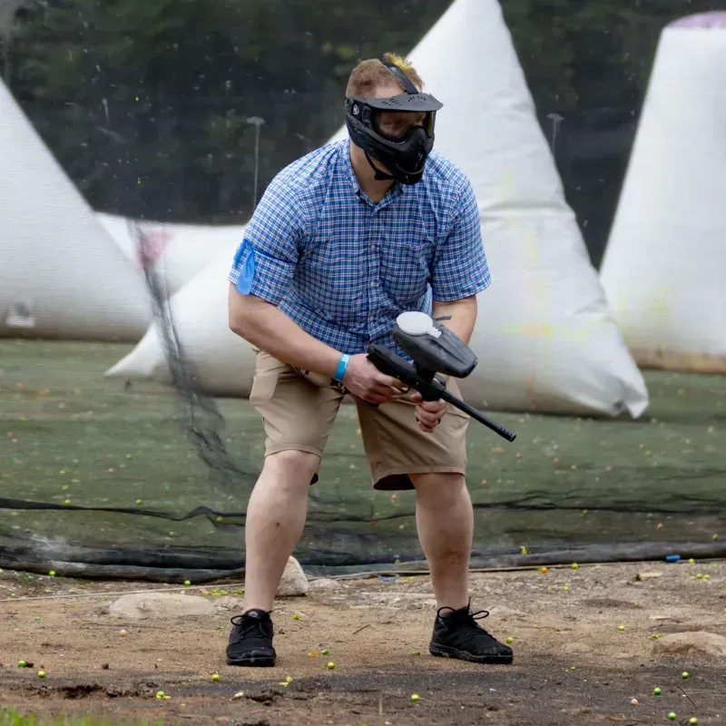Man in paintball mask aiming a paintball gun outdoors. He's wearing a blue shirt, shorts, and black shoes.