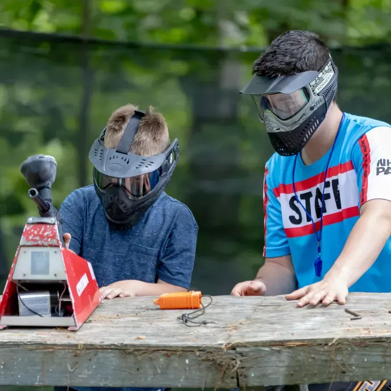 Two boys in paintball masks check equipment at an outdoor table. One wears a staff shirt.