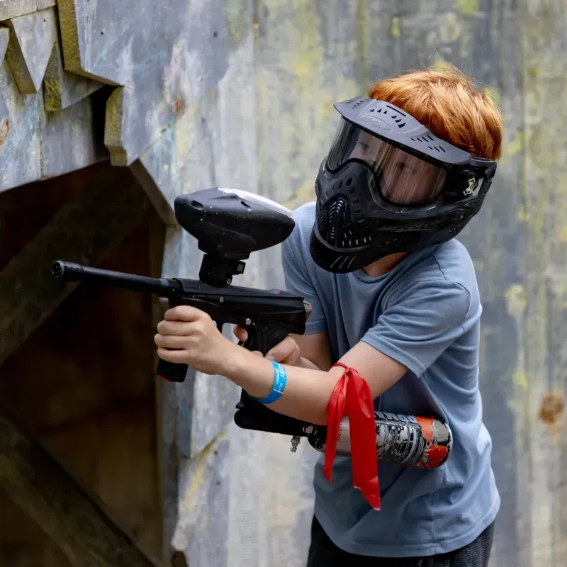 Boy with red hair in paintball gear aiming a paintball gun.