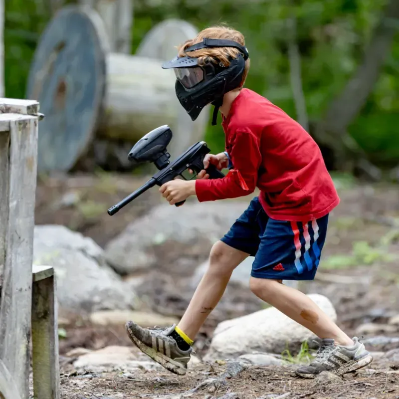 Boy in red shirt and mask plays paintball outdoors, aiming gun.