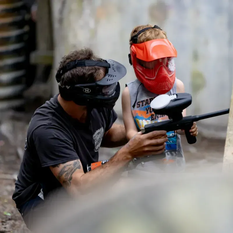 Man helping a child with a paintball gun; both wear protective masks in an outdoor setting.