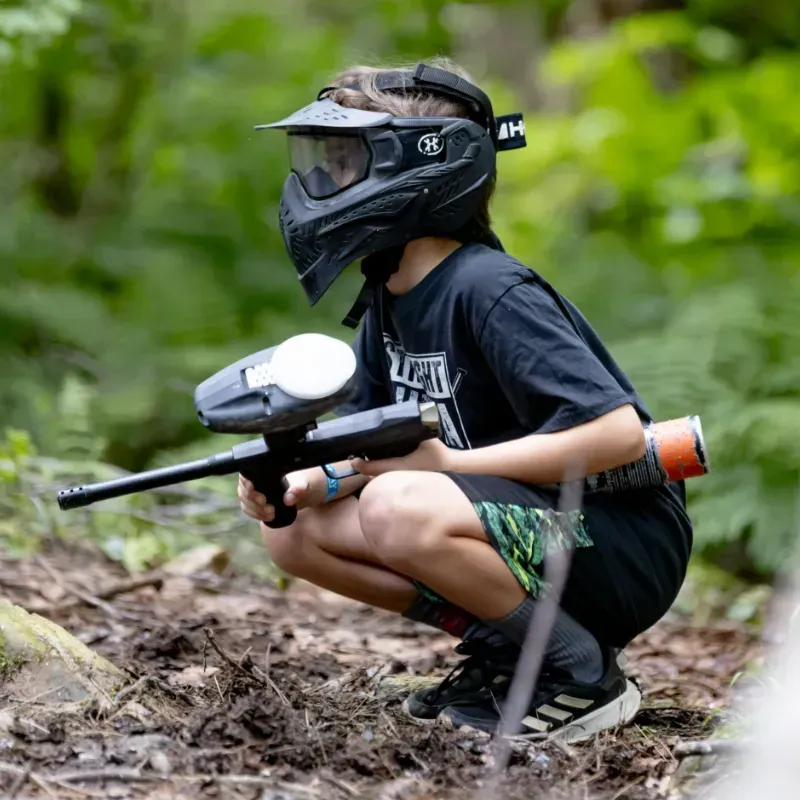 A young person in paintball gear crouches in a forest, aiming their paintball gun.