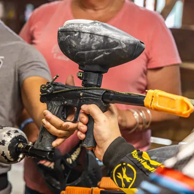 Person holding a black paintball gun with orange barrel cover; others nearby.