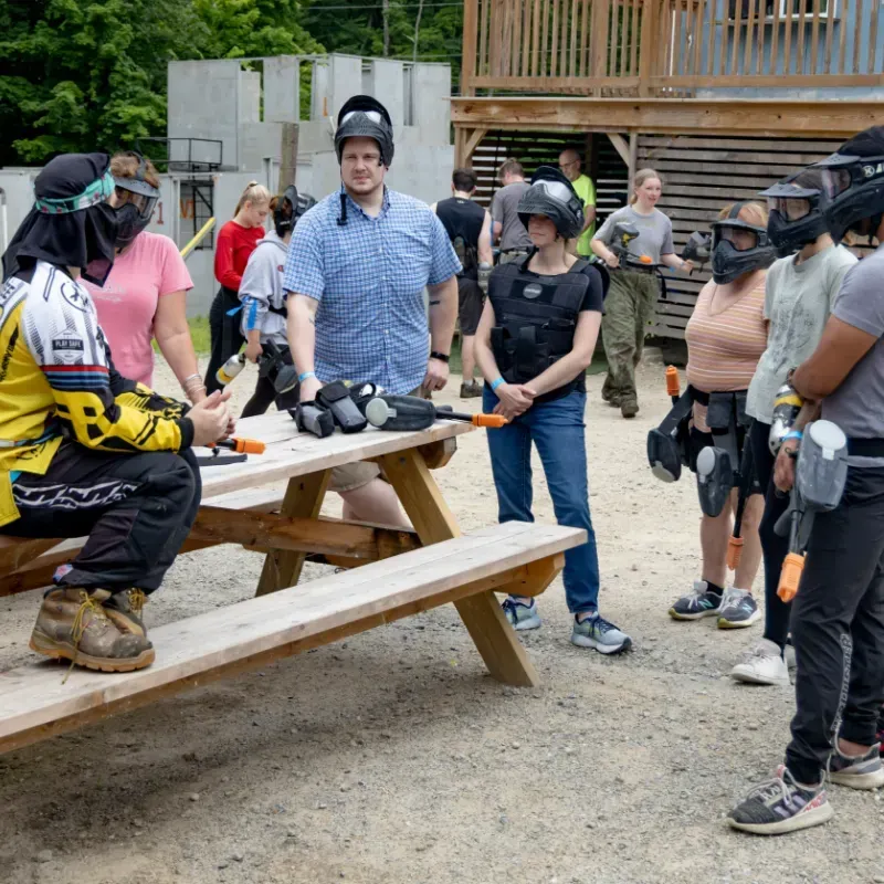 People in paintball gear at a course. Some sit on a bench, others stand with markers. Outdoor setting.