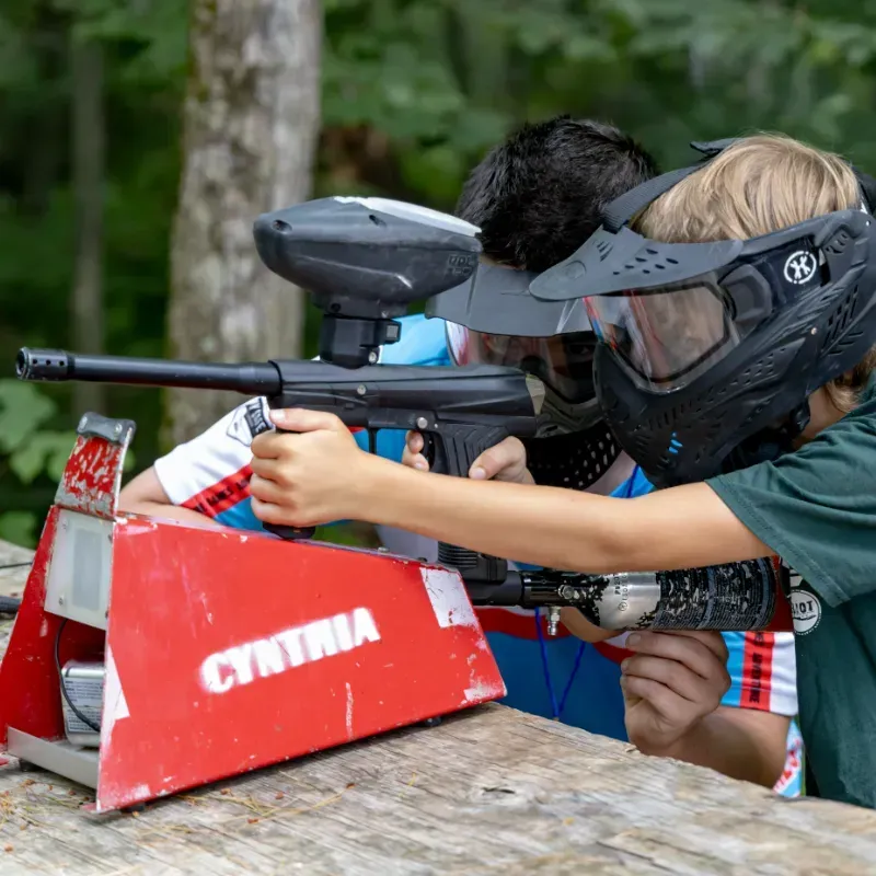 Two children playing paintball, aiming guns behind a red shelter in a wooded area.