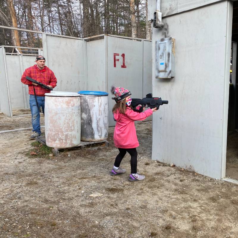 A young girl in pink aims a toy gun in an outdoor arena. An adult man stands nearby.