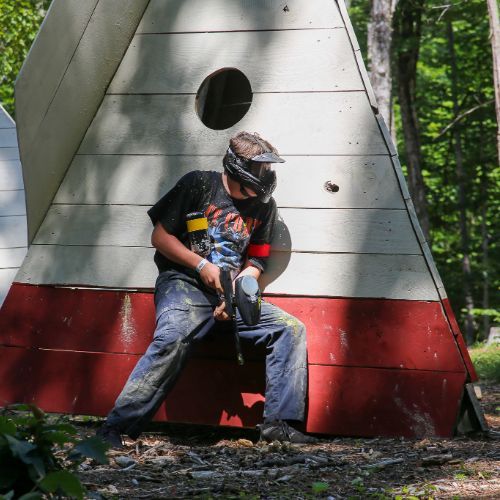 A person in paintball gear aiming a marker, standing near a wooden structure in a wooded area.