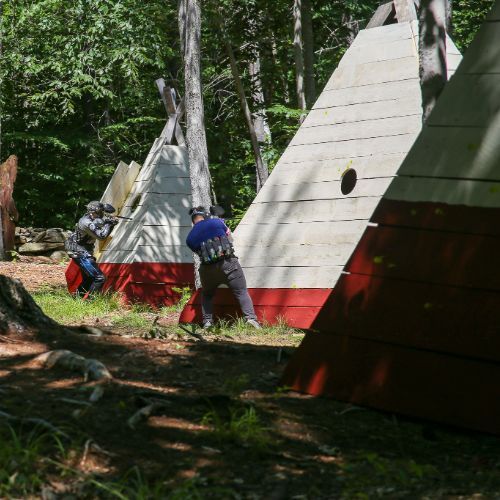 People playing paintball behind red and white triangular wooden structures in a forest setting.