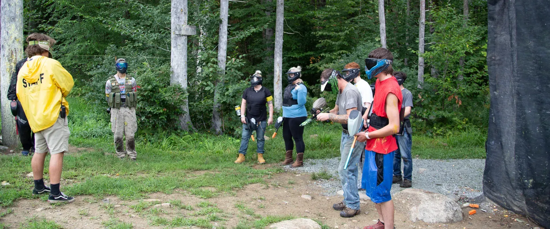 People in paintball gear, some with masks on, stand in a wooded area.