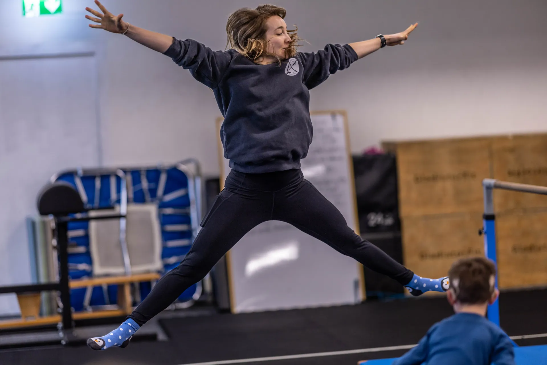 A person performs a star jump in an indoor gym in Thalwil with a child watching in the foreground.