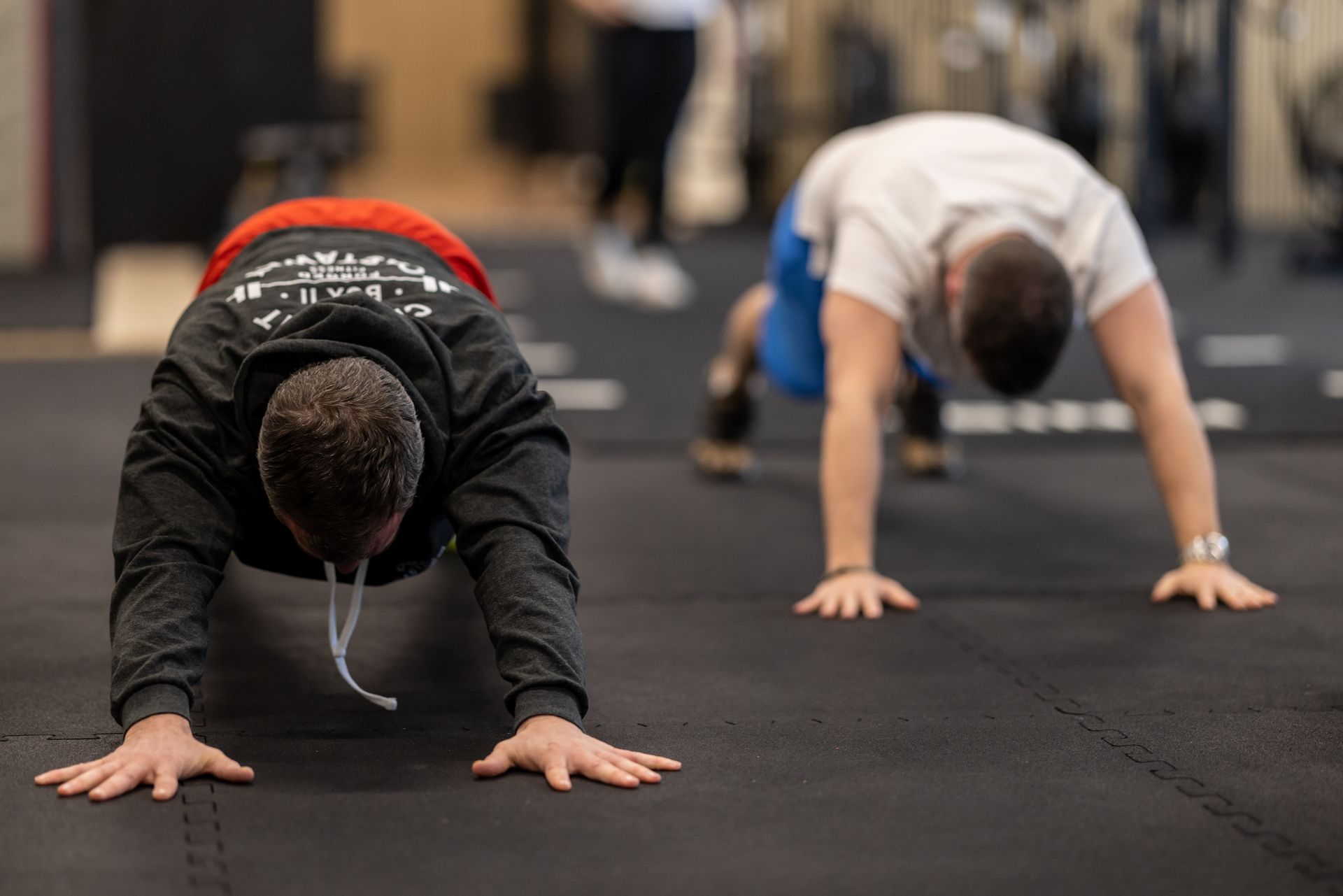 Two people in a gym in Thalwil perform plank exercises on black floor mats.