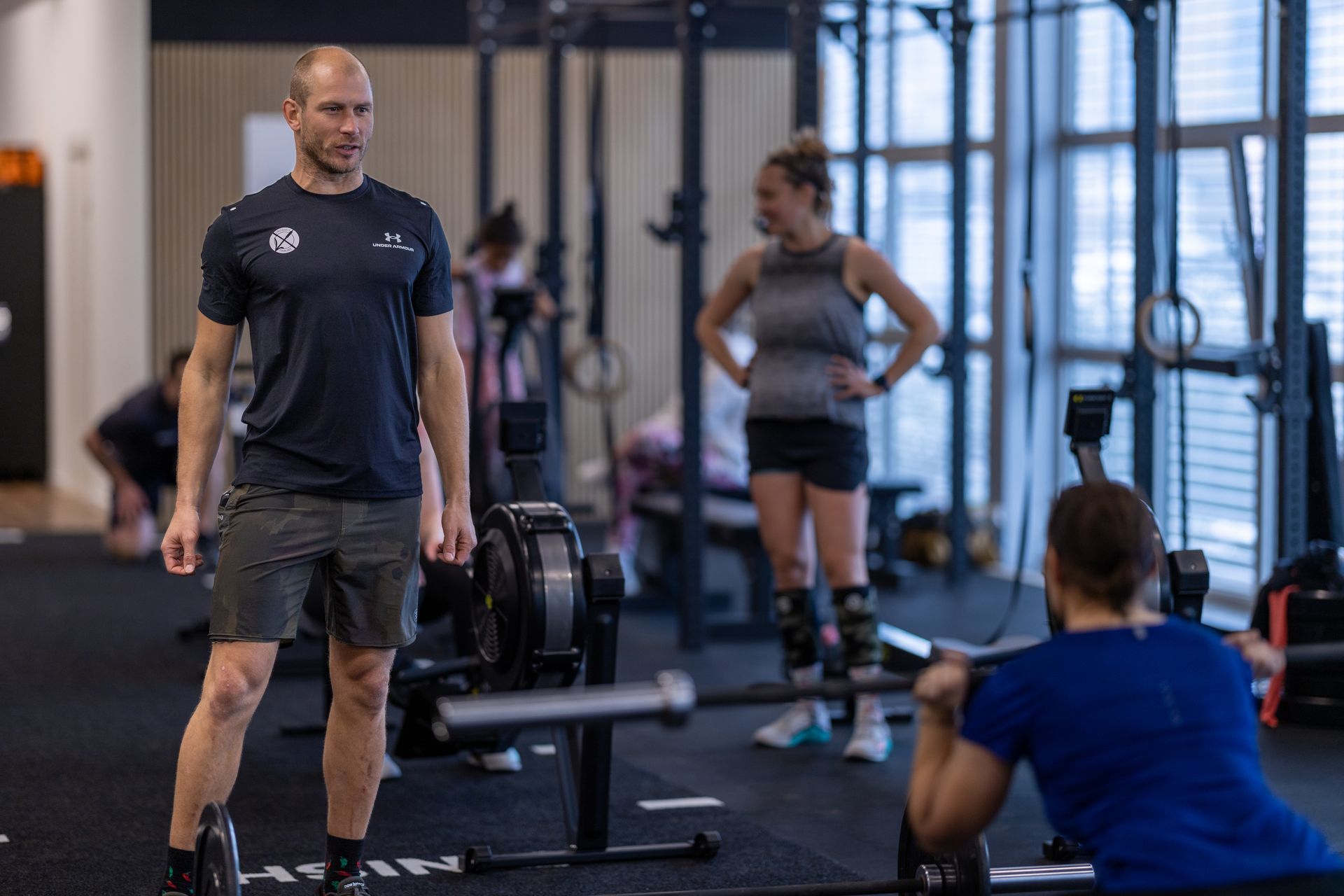 A coach stands in a gym in Thalwil, watching someone squat with a barbell while another person stands nearby