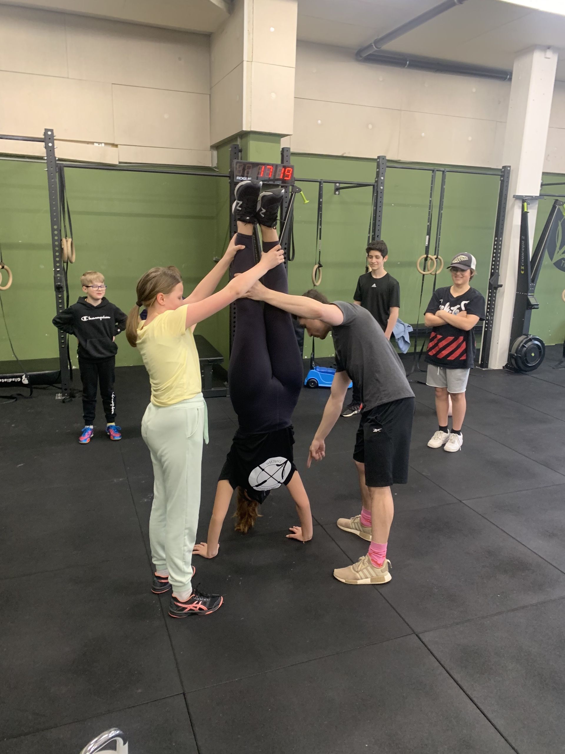 A person in a gym in Thalwil performs a handstand while two people assist by holding their legs upright.