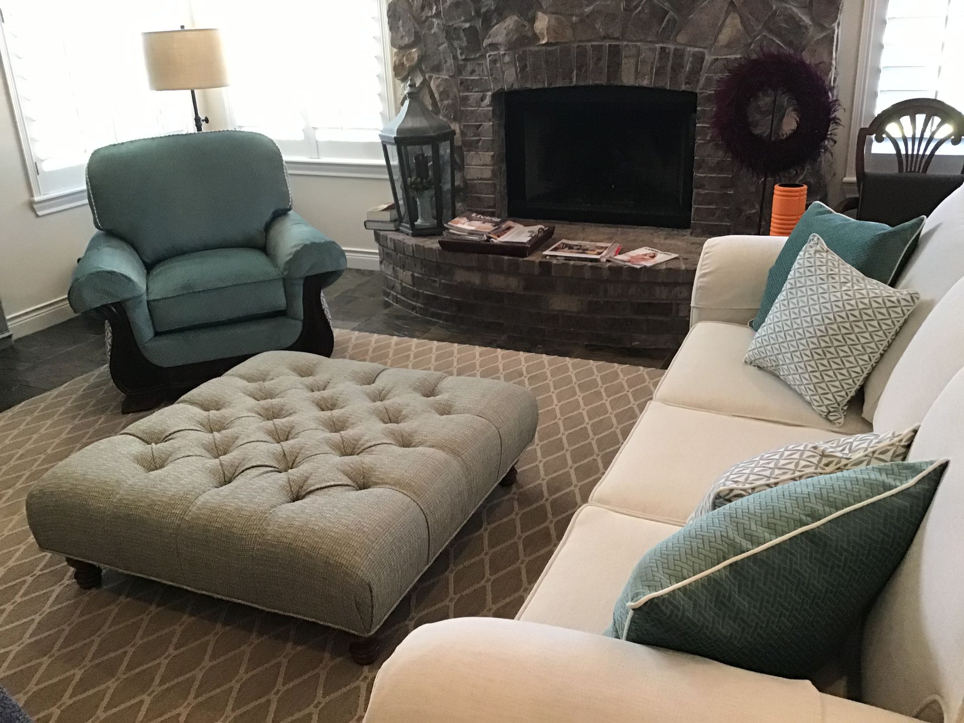 A living room with a beige sofa, blue armchair, and large tufted ottoman on a patterned rug in front of a stone fireplace.