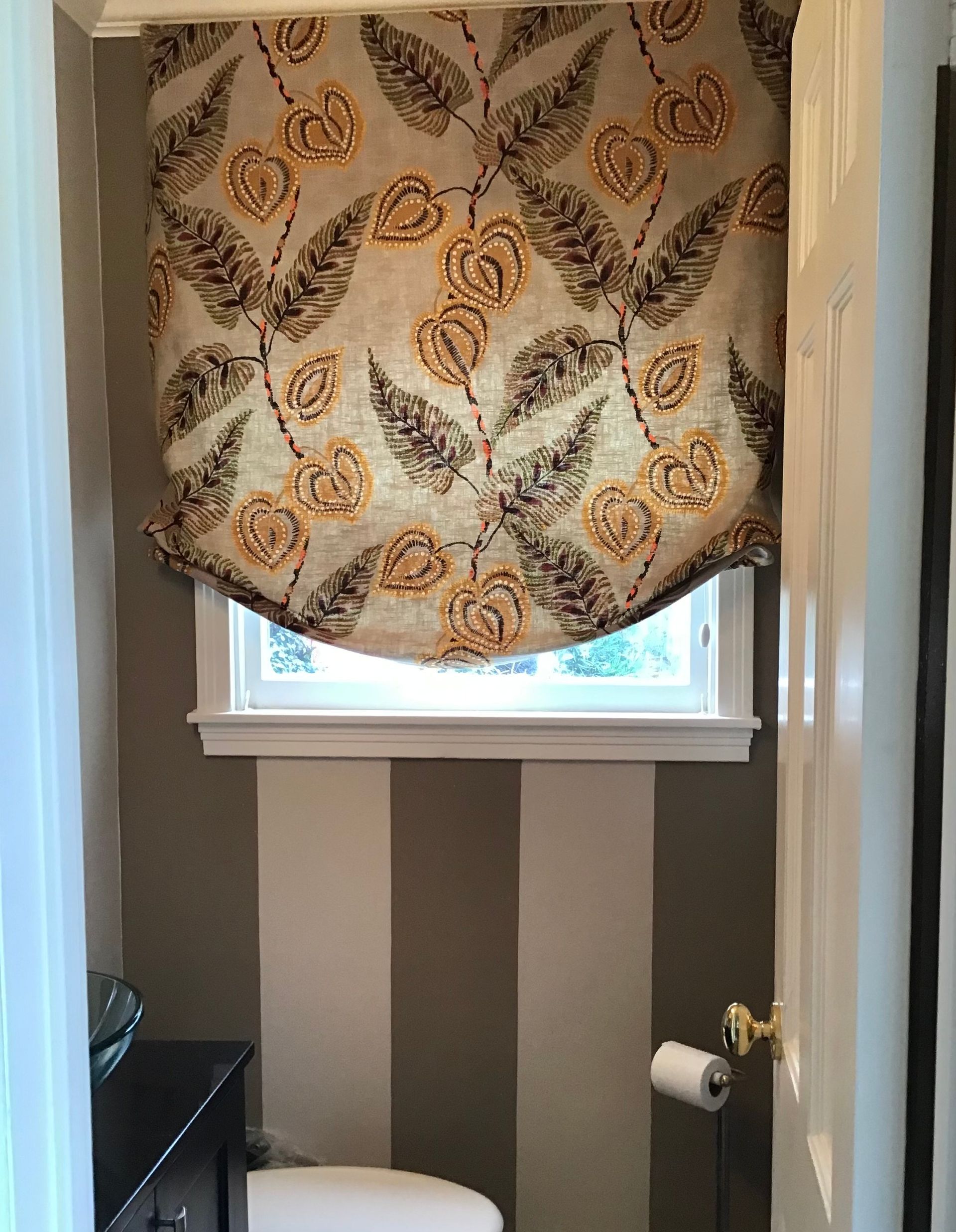 A powder room with vertically striped brown and beige walls, a small window with a floral Roman shade, and a toilet.