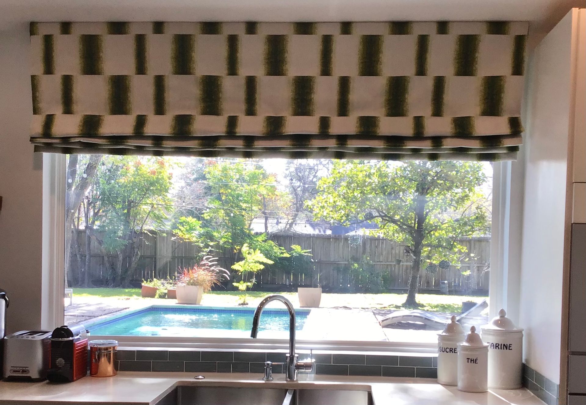 A kitchen window overlooking a backyard swimming pool, featuring a fabric Roman shade with a green and white geometric print.