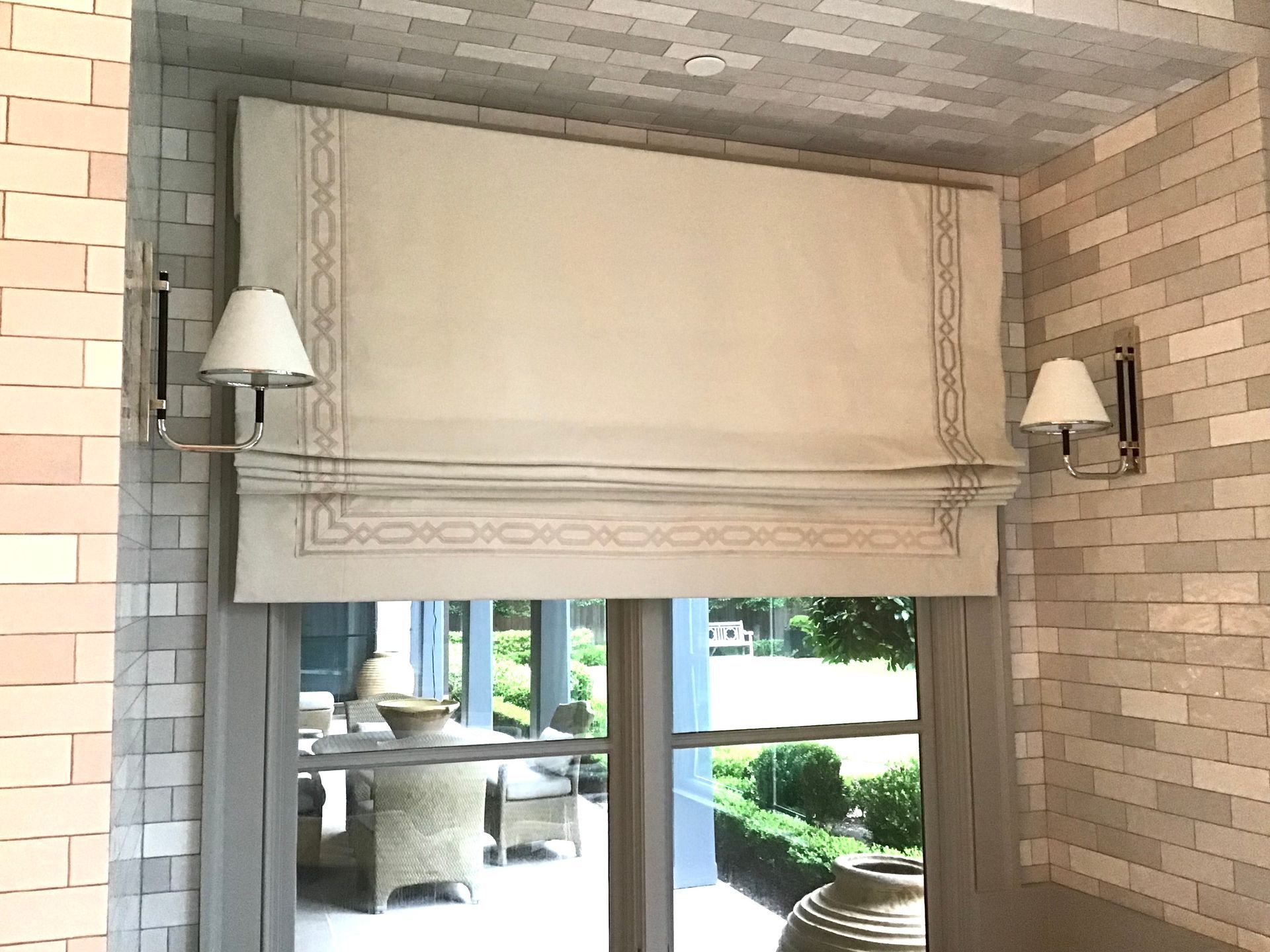 A fabric Roman shade with a decorative border hanging in a window between two wall sconces on a light-colored brick wall.
