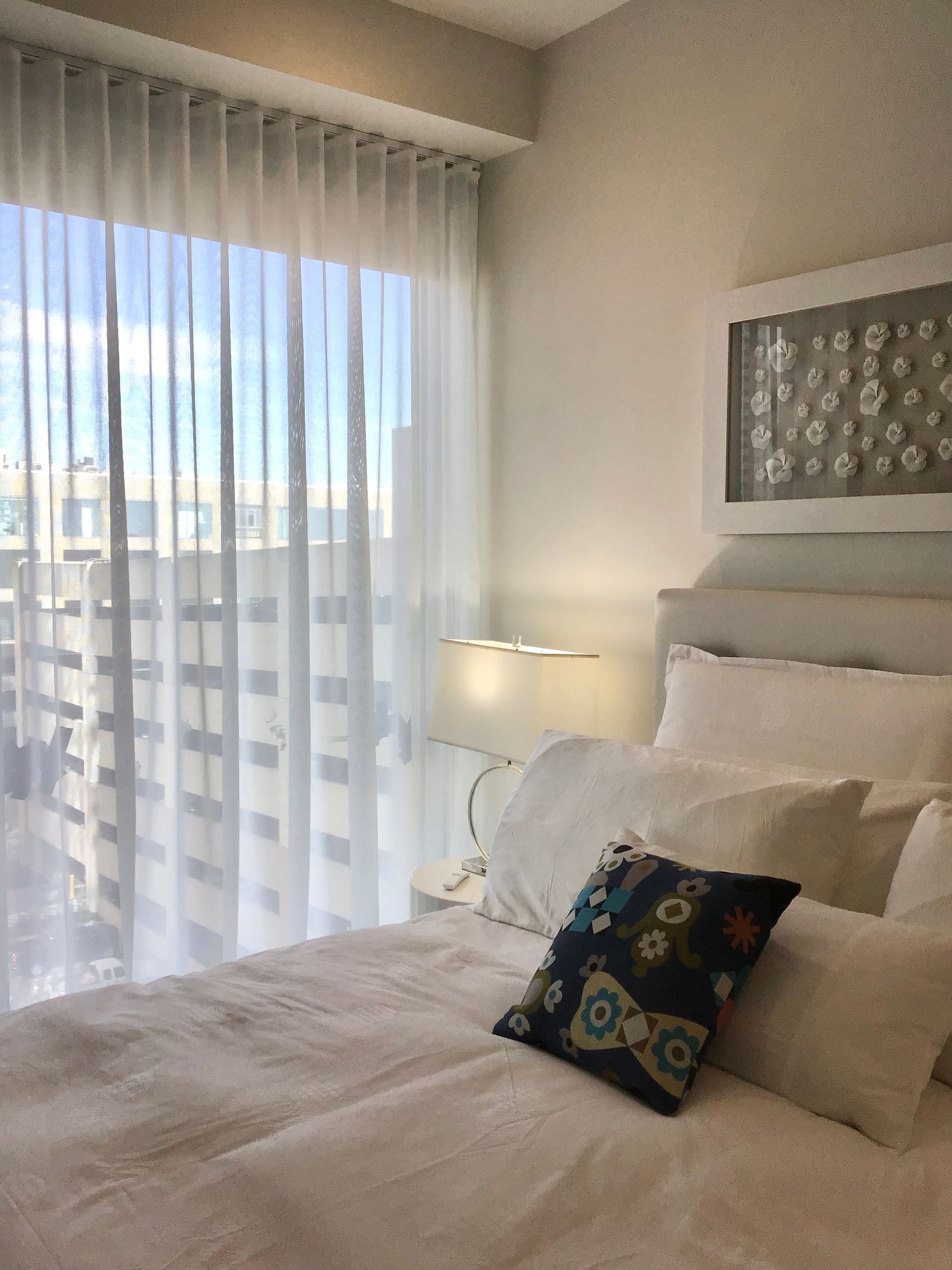 A sunlit bedroom features a neatly made bed with white bedding, a decorative dark accent pillow, and a soft bedside lamp.