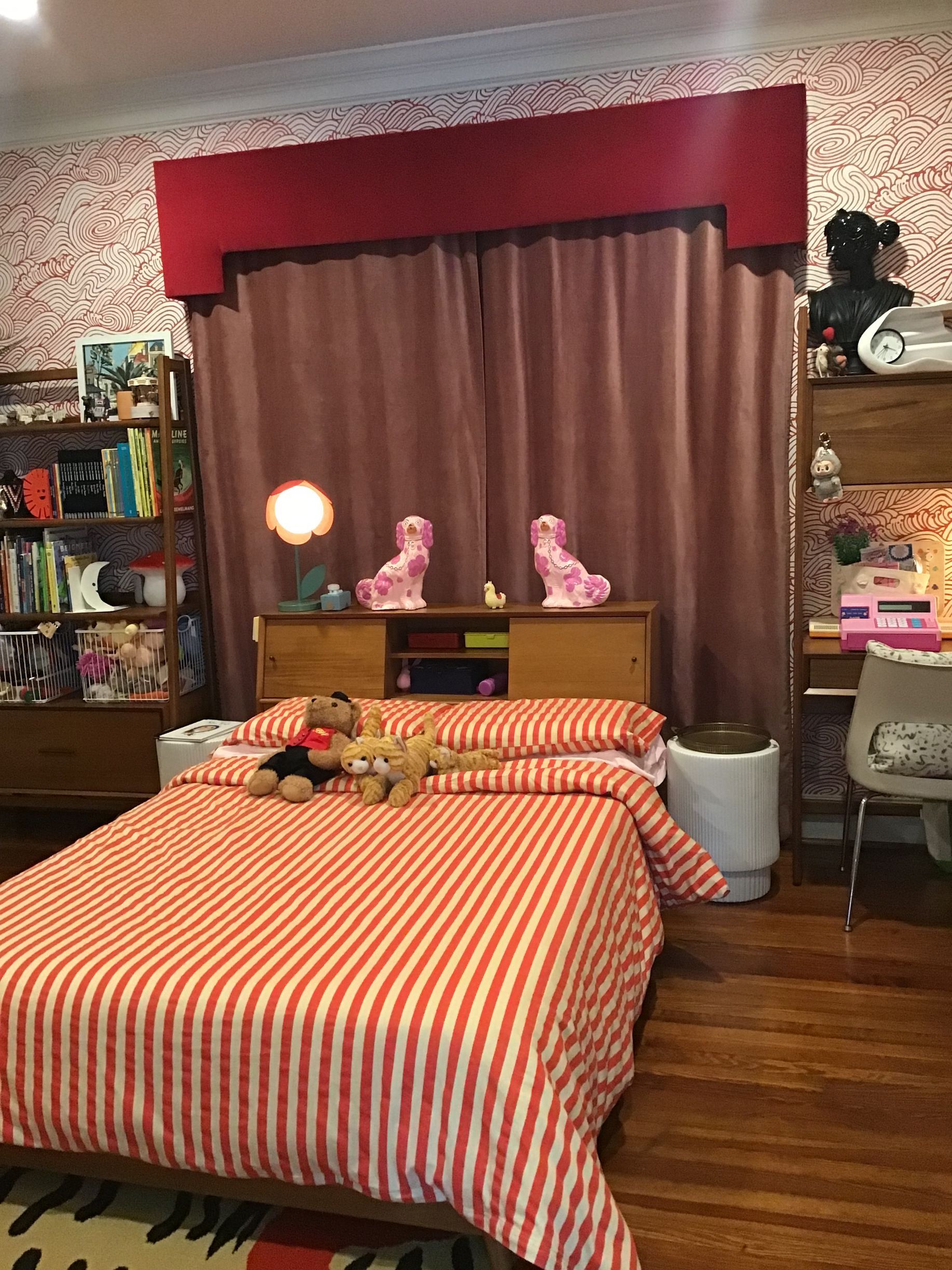 A bedroom with a bed featuring orange-and-white striped bedding, wood furniture, patterned wallpaper, and a red valance.