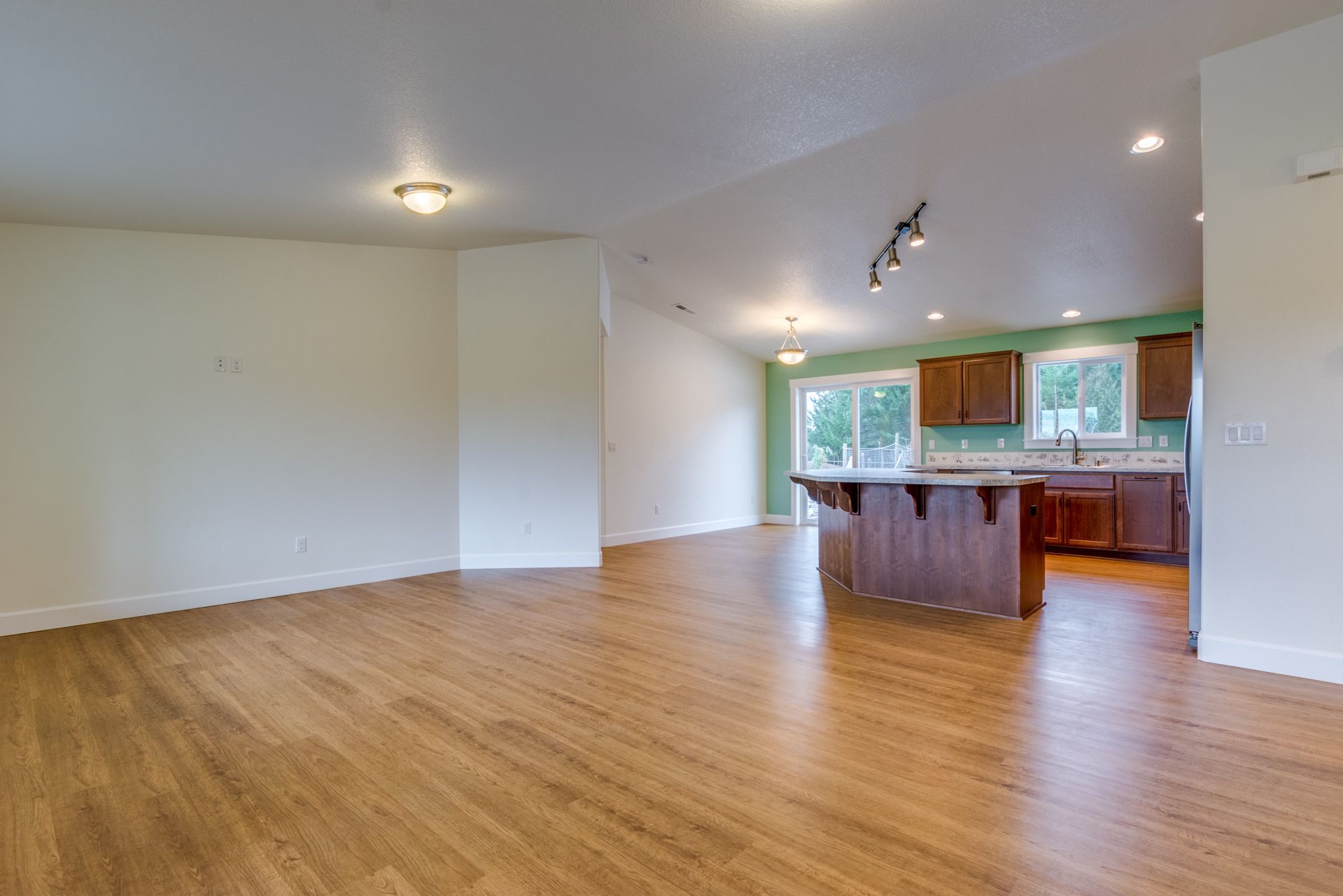 An empty living room with hardwood floors and a kitchen in the background.