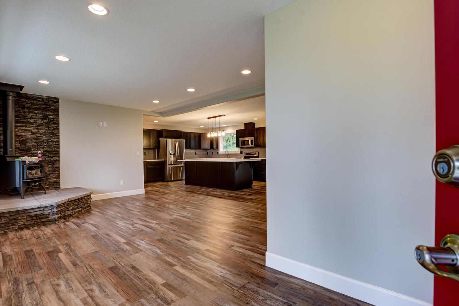 A red door is open to a living room with hardwood floors and a kitchen.