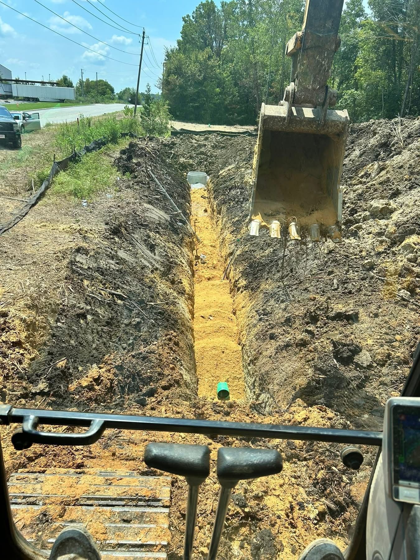 Excavator digging a deep trench for water, sewer, or drainage system installation near a roadway.