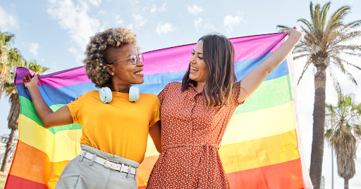 Two smiling individuals holding a rainbow Pride flag outdoors, representing LGBTQ+ inclusive and aff