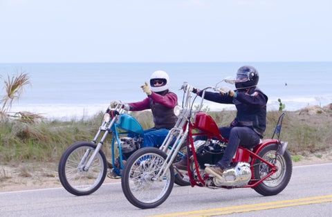 Two people are riding motorcycles down a road near the ocean.