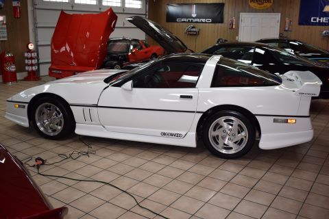 A white corvette with the hood up is parked in a garage.
