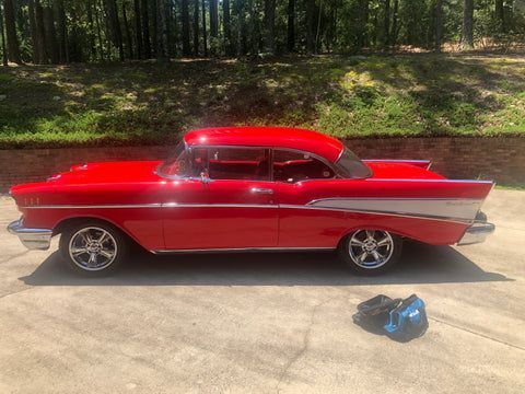 A red classic car is parked in a driveway next to a brick wall.