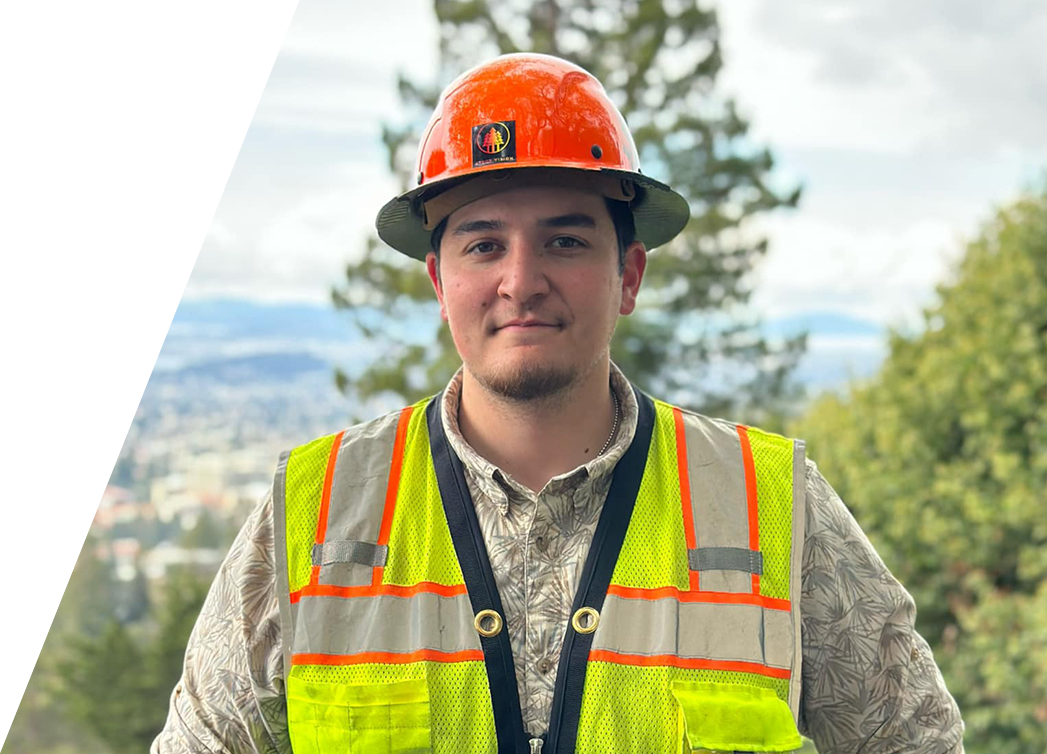 Arbor Vision member wearing an orange hard hat and reflective safety vest stands in front of a scenic backdrop with trees and a distant city view.