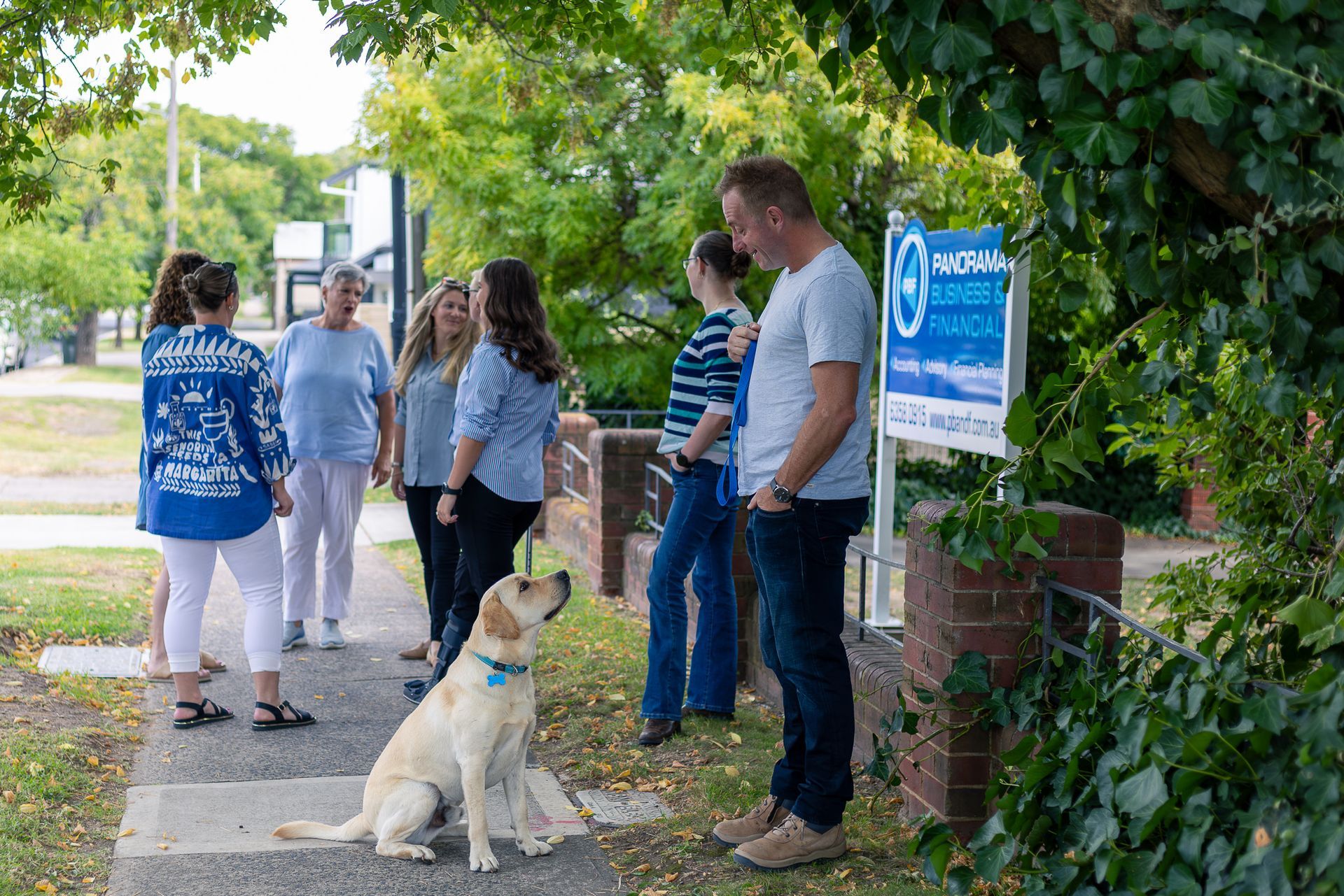 Group standing near a campus sign with a yellow dog sitting on the sidewalk under trees