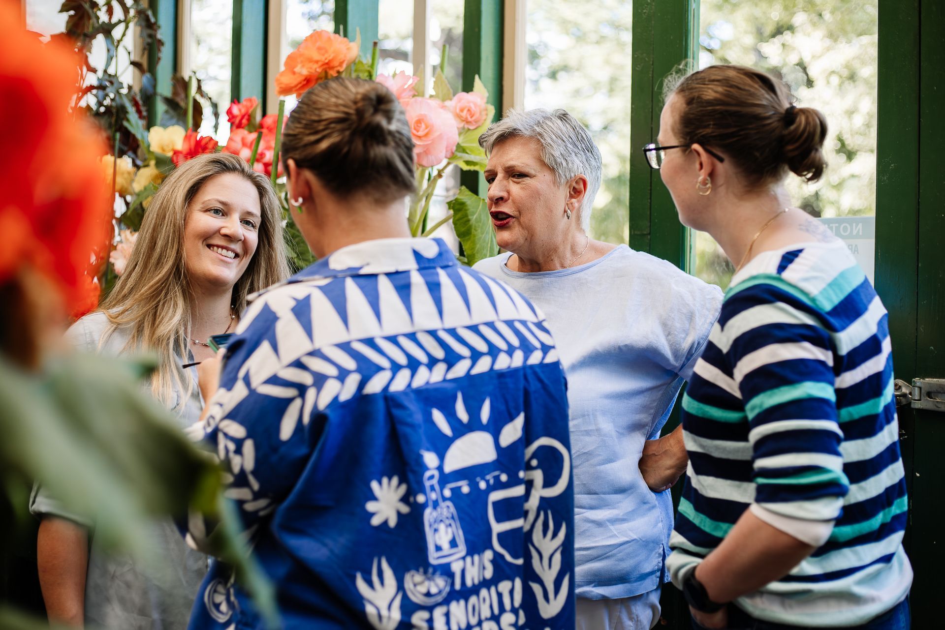 Group of people chatting outdoors, including one in a blue patterned shirt, in a bright garden setting