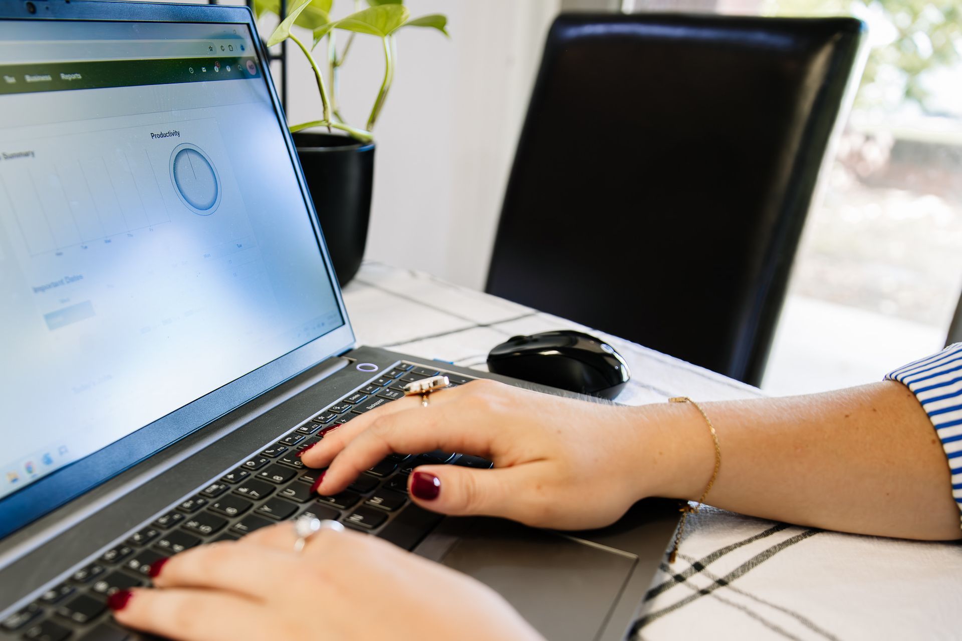 Person typing on a laptop at a table beside a plant and chair in a bright room